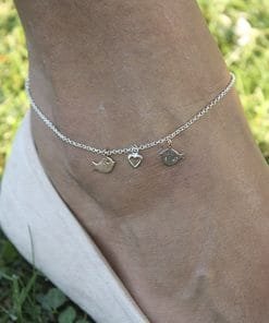 Gül Kuş ve Kalp İncirlü Taşsız Halhal giyen bir ayak bileğinin yakın çekimi, iki kuş takısı ve bir kalp takısı, çim ve açık renkli bir ayakkabıya karşı.